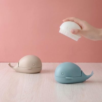 Brosse design baleine blanche tenue en main au-dessus de deux baleines beige et bleue